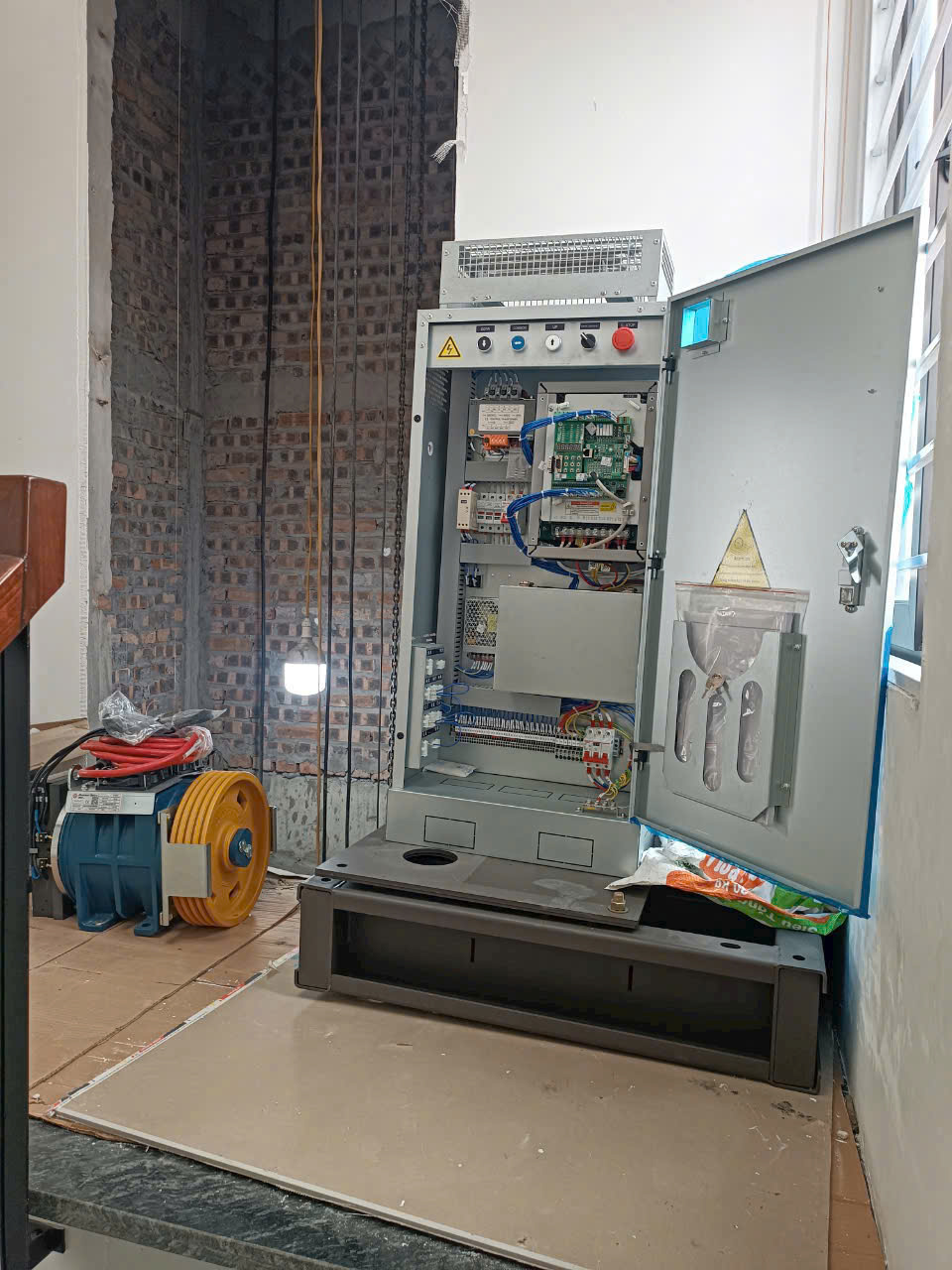 Tủ điều khiển thang máy đang lắp đặt cạnh hố thang và động cơ kéo màu xanh vàng